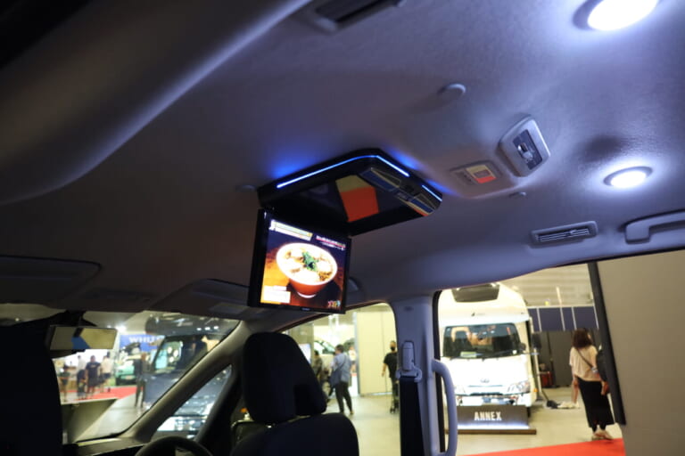 展示車の後席にはオプションのフリップダウンモニター（10万5000円•税込）を搭載。ほかにも18L冷凍冷蔵庫やルーフエアコン、200W薄型ソーラーパネルなどクルマ旅を快適にするオプション装備が用意されている。｜思い切った「“5人乗り”コンセプトミニバン」で自由な旅！独自アルミフレーム構造で“十分な収納力”＆テーブル・テレビも装備！サブバッテリーなど電装類も良い感じ！