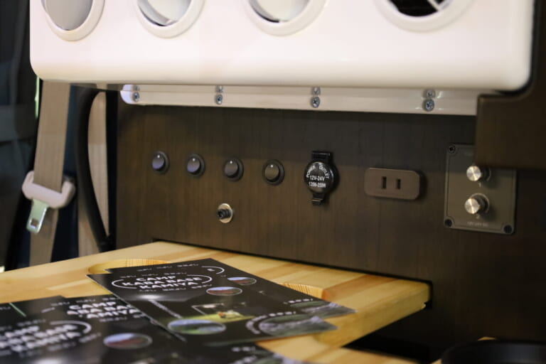 クーラー下には照明のスイッチやAC100Vコンセントなどを配置。外部電源接続時に使用できる。｜「えぇーっ 流石にスゴイ」「本当に軽自動車ですか？」バンライファーにぶっ刺さる！まさかの天然木が胸を打つ、あれもこれも詰まった軽キャンパー。