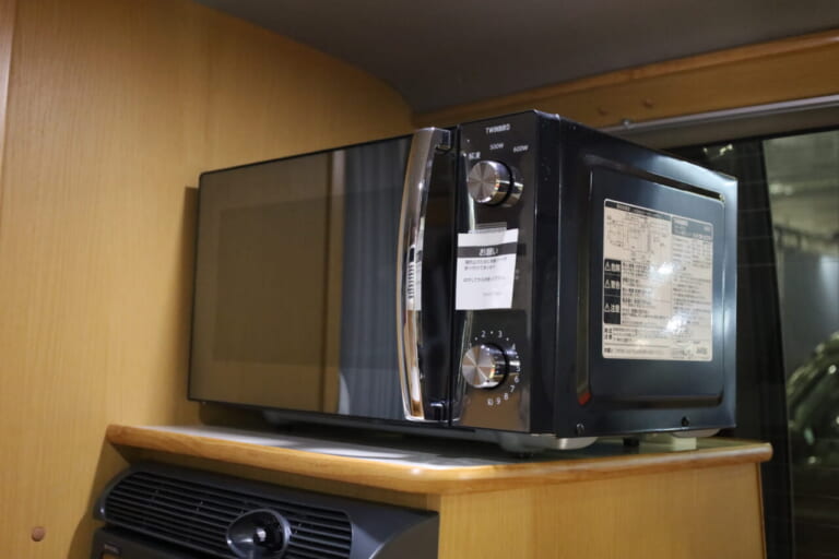 車内調理で活躍する電子レンジを装備。電子レンジの下には40Lの横開き冷蔵庫を標準装備。庫内から出したものをすぐに調理できる。ともに標準装備品だ。｜「おいおい、これは住めちゃうぞ」クルマの中に電子レンジや冷蔵庫、キッチンまで装備。もはや1Kの部屋みたいで便利に使えそう。