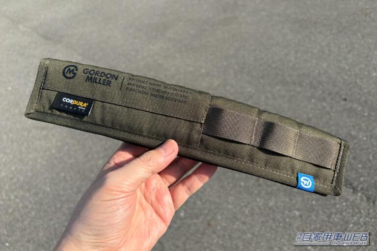 オートバックスのオリジナルブランド、ゴードンミラーのコーデュラ シートベルトパッド(OLIVE DRABカラー)を手に持っている写真|「シートベルトって洗えないよね…」夏場は汗でニオイも気になる。だったらコレ使ってみて! デザイン性が高く通気性も抜群「ゴードンミラー・コーデュラ シートベルトパッド」